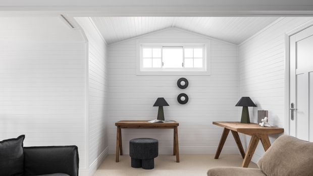 A bright, compact white-paneled room styled as a minimalist home office or study. It features two timber desks, dark table lamps, a small round stool, a black sofa and neutral armchair, a high horizontal window, white tongue-and-groove wall and ceiling lining, and pale carpet. The pitched ceiling and restrained black-and-white palette create a clean, calm atmosphere.