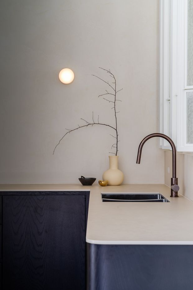 A refined minimalist kitchen vignette featuring a pale beige stone or engineered-stone benchtop, dark timber cabinetry, an inset stainless-steel sink, and a curved dark bronze or brown tap. A warm wall light, sculptural ceramic vase with a bare branch, and small ceramic vessels add understated decorative detail. A white-framed window provides natural light on the right.