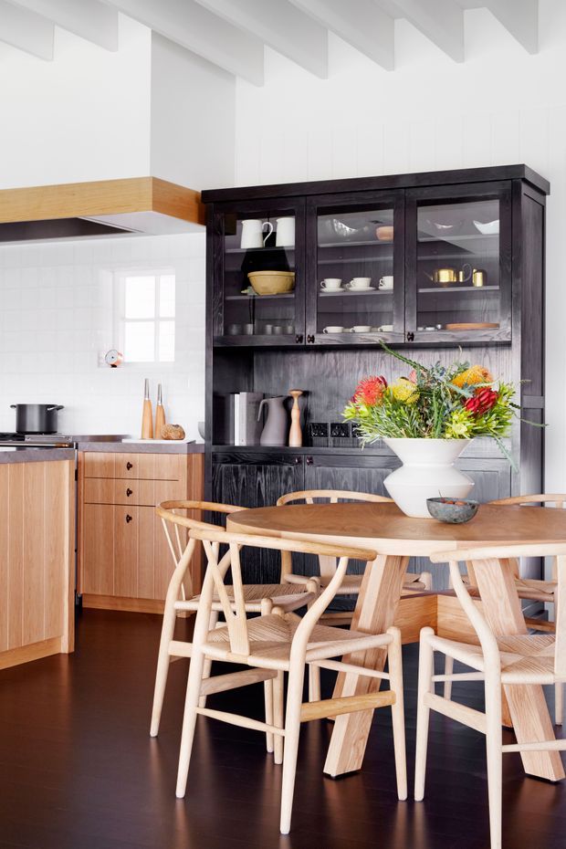 Bright, refined kitchen-dining interior with white tiled walls and exposed white ceiling beams. A warm timber kitchen with timber rangehood, dark timber flooring, and a large black stained timber glazed display cabinet contrasts with light timber dining furniture. The dining table is styled with a large colourful floral arrangement.