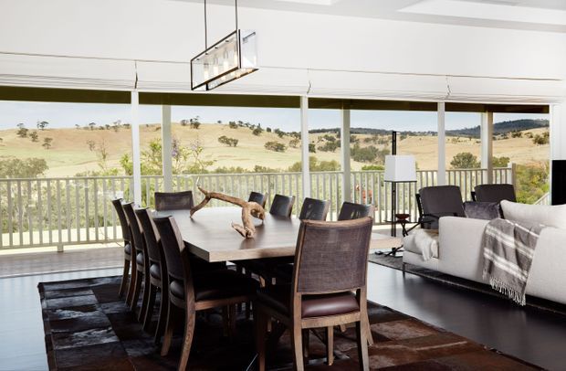 A bright, spacious open-plan dining and living area with expansive floor-to-ceiling glazing and views across rolling rural hills. A large dark timber dining table with matching high-back chairs sits on a patchwork hide rug, while a light upholstered sofa and armchair occupy the adjacent sitting area. The room features a white coffered ceiling, dark flooring, a contemporary rectangular pendant light, and a full-width covered veranda with balustrade visible beyond the glazing.