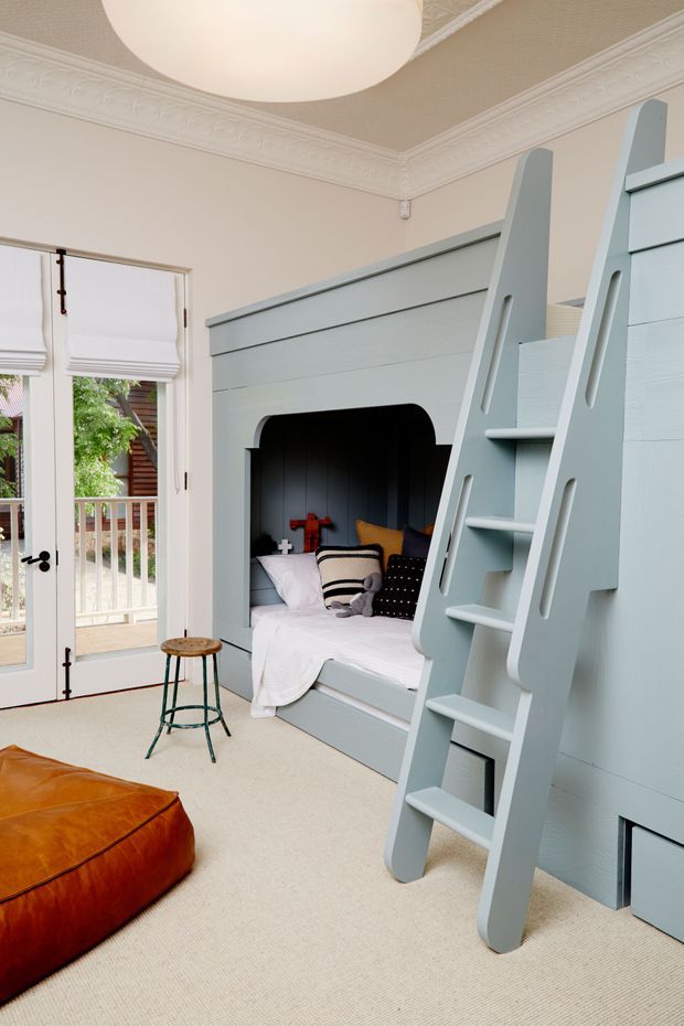 A bright, characterful bedroom featuring a custom pale blue built-in bunk bed with an integrated ladder and a lower sleeping nook. The room has ornate white ceiling cornices, pale walls, French doors with white blinds opening to a balcony, a large white pendant light, neutral carpet, and a tan leather ottoman. Decorative pillows and bedding add small dark and warm accents.