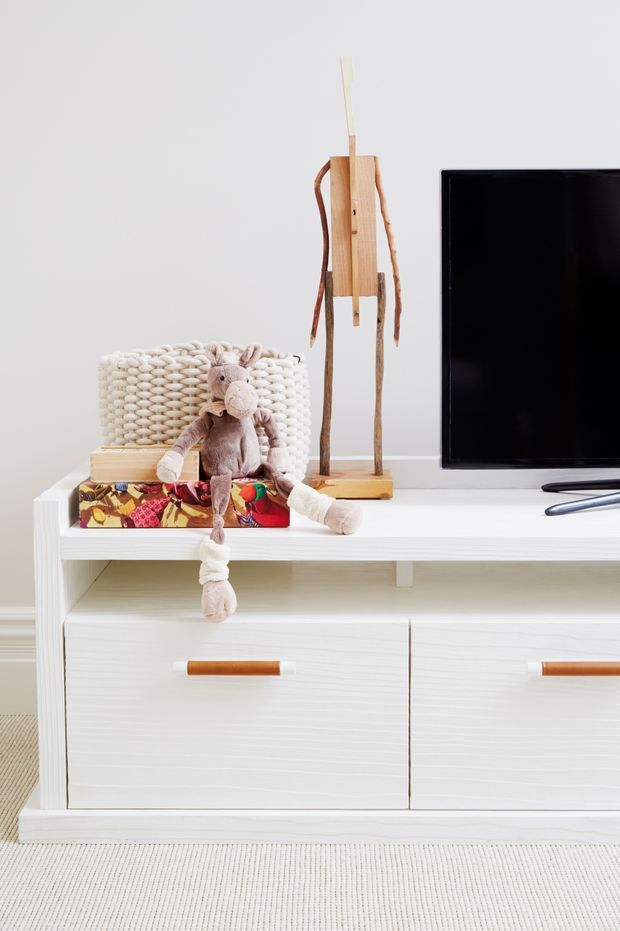 A bright, minimalist living room vignette featuring a low white timber media console with drawers, a flat-screen television, a woven basket, a wooden decorative figure, wrapped gift boxes, and a plush stuffed toy. The scene uses a soft neutral palette with warm timber accents and textured woven details.