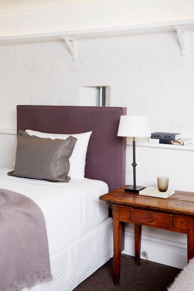 A warmly styled bedroom featuring a white textured plaster wall with a long built-in shelf, a deep mauve upholstered headboard, white bedding, taupe cushions and throw, and a rustic dark timber bedside table with a lamp and books. The room has dark carpet and traditional wall detailing, creating a refined, cozy atmosphere.