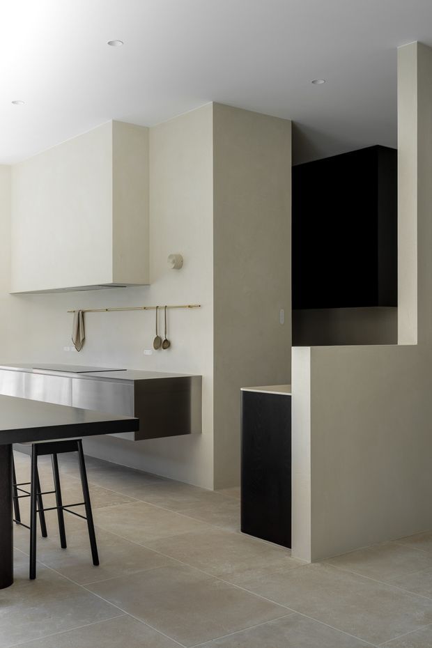 A minimalist contemporary kitchen with pale plastered walls, large neutral floor tiles, stainless-steel lower cabinetry, a dark island benchtop, and a concealed dark pantry or utility zone behind a partial-height partition. A slim brass rail with hanging utensils adds a small accent, while recessed ceiling lights and clean geometric forms create a restrained, modern interior.