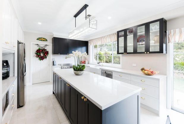 Bright, spacious contemporary kitchen with a large white island, dark charcoal cabinetry, white perimeter cabinetry, glass-front upper cabinets, stainless-steel appliances, and a linear pendant light. The room has abundant natural light, pale tiled flooring, and garden views through the windows and glazed door.