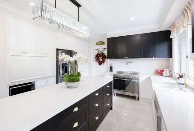 Bright, spacious contemporary kitchen with a large white island featuring dark built-in cabinetry, white perimeter cabinetry, pale tiled flooring, and black upper cabinets. A stainless-steel range cooker, integrated appliances, large windows with timber blinds, and a linear crystal pendant light complete the refined high-contrast scheme.