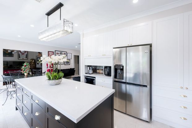Bright, spacious contemporary kitchen with a large white island, dark painted cabinetry, extensive white built-in cabinetry, stainless-steel refrigerator, integrated appliances, and a linear glass pendant light. A dining and living area is visible beyond the kitchen, with festive decor and artwork.