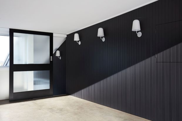 A minimalist interior hallway or entry space with a dramatic black vertically panelled feature wall, four white wall sconces, a black-framed glazed door, and a pale polished concrete floor. Strong natural light creates a diagonal shadow across the wall.