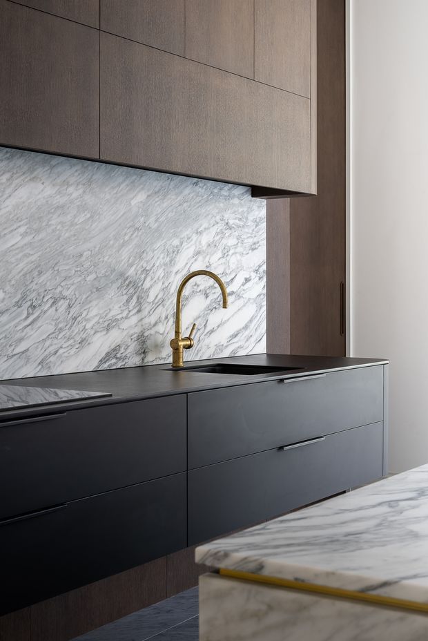 A refined contemporary kitchen with dark matte lower cabinetry, warm wood-toned overhead and tall joinery, a dramatic white-and-grey marble splashback and island surface, and a brass gooseneck tap. The compact composition highlights integrated storage, a dark benchtop and sink, and a luxurious neutral material palette.