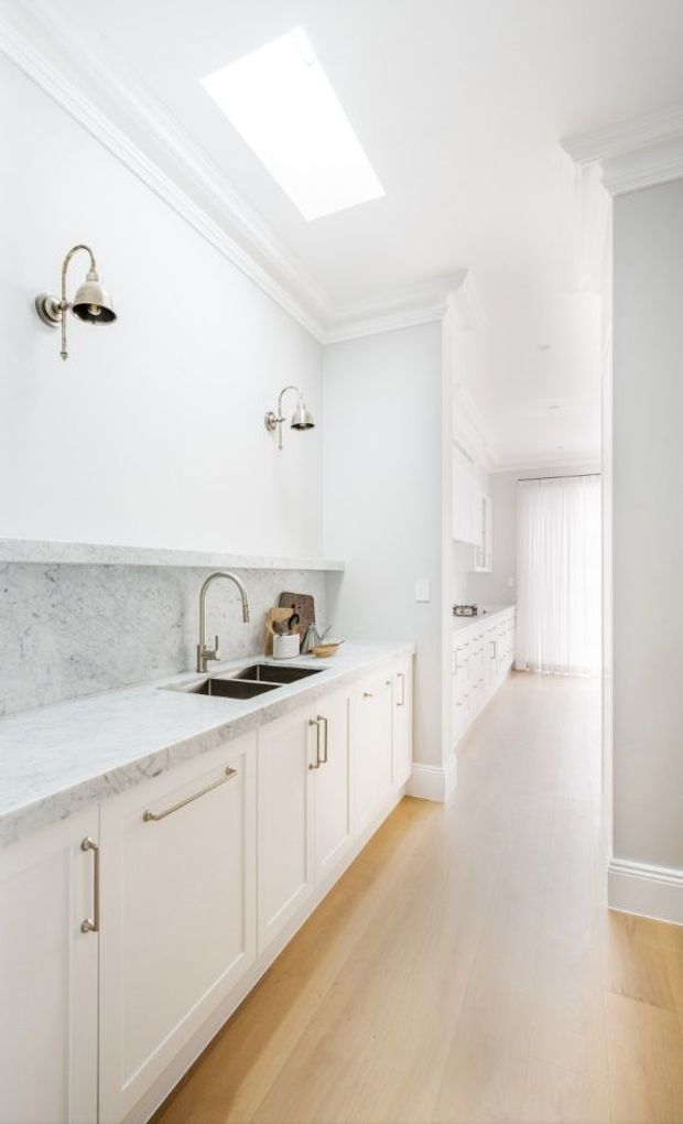 A bright, narrow galley-style kitchen with white raised-panel cabinetry, pale veined stone benchtops and backsplash, a double undermount sink with a curved metal tap, and brass-toned wall sconces. Light timber flooring, white walls, ornate crown moulding, and a large skylight create an airy, classic interior. The kitchen extends toward a bright adjoining area with additional cabinetry.