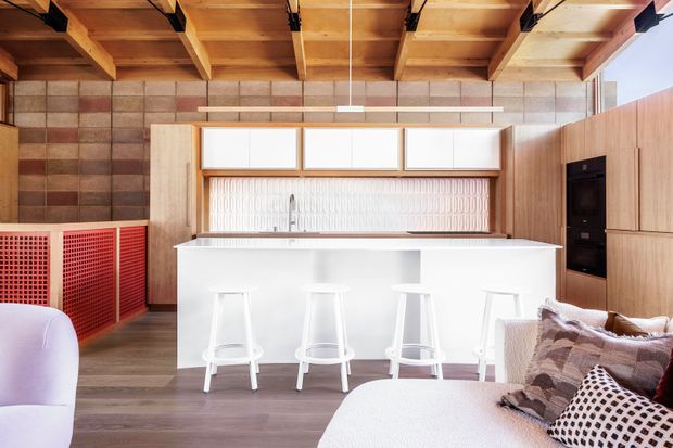 Bright open-plan kitchen and living area featuring a white island with four stools, pale timber cabinetry, a ribbed backsplash, exposed timber ceiling beams, and a textured masonry feature wall. A light sofa with patterned cushions occupies the foreground, while red perforated built-in screening adds a bold accent.
