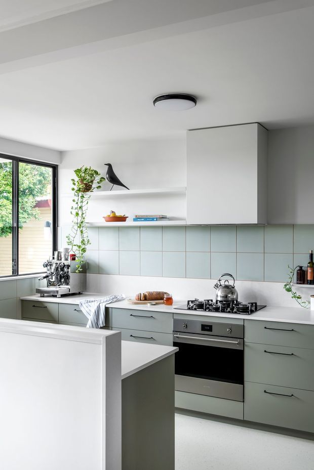 Bright, modern kitchen with pale sage-green lower cabinetry, white benchtops, and a light blue-green tiled splashback. The space includes a freestanding stainless-steel oven and gas cooktop, kettle, espresso machine, open display shelving, and a compact peninsula. Large black-framed glazing brings in daylight and views of greenery. Minimal decorative styling includes a trailing plant and black bird ornament.
