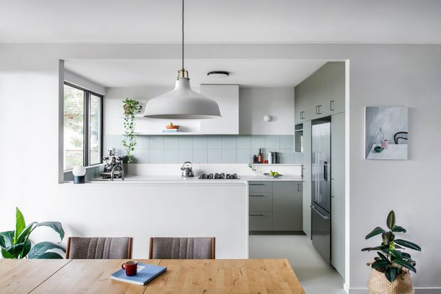 Bright contemporary open-plan kitchen and dining area with a white peninsula, pale grey-green cabinetry, pale blue-grey tiled splashback, stainless-steel appliances, timber dining table, upholstered dining chairs, indoor plants, and a large sculptural pendant light. The kitchen is framed by a wide opening and adjacent full-height glazing, creating an airy, clean setting.