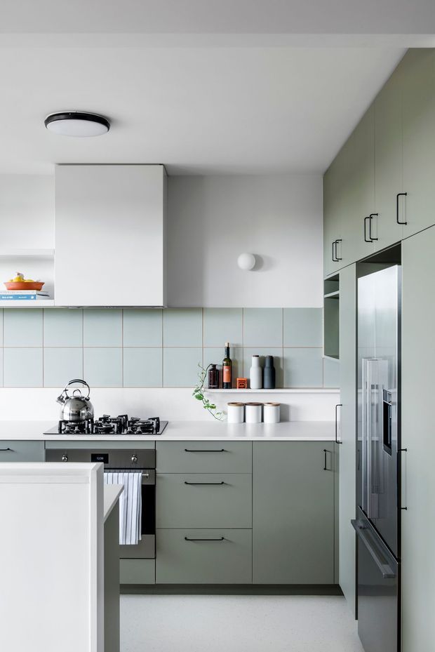 A compact, contemporary kitchen with pale sage-grey cabinetry, white benchtops, and a grid of pale blue-grey square tiles forming the splashback. The kitchen includes a gas cooktop and oven, stainless-steel kettle, integrated refrigerator, open shelving, and a small peninsula or island in the foreground. Minimal black hardware and simple ceiling and wall lighting contribute to a clean, restrained aesthetic.