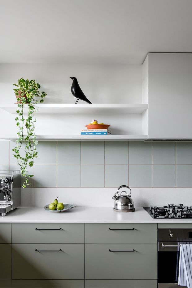 A clean, compact contemporary kitchen with pale grey-green flat-panel cabinetry, a white benchtop, and a tiled splashback in muted grey-blue with fine contrasting grout lines. Open white shelving displays a trailing plant, black bird sculpture, and a small stack of books. The kitchen includes a stainless-steel kettle, gas cooktop, built-in oven, and coffee machine, with soft natural light creating a calm, refined atmosphere.