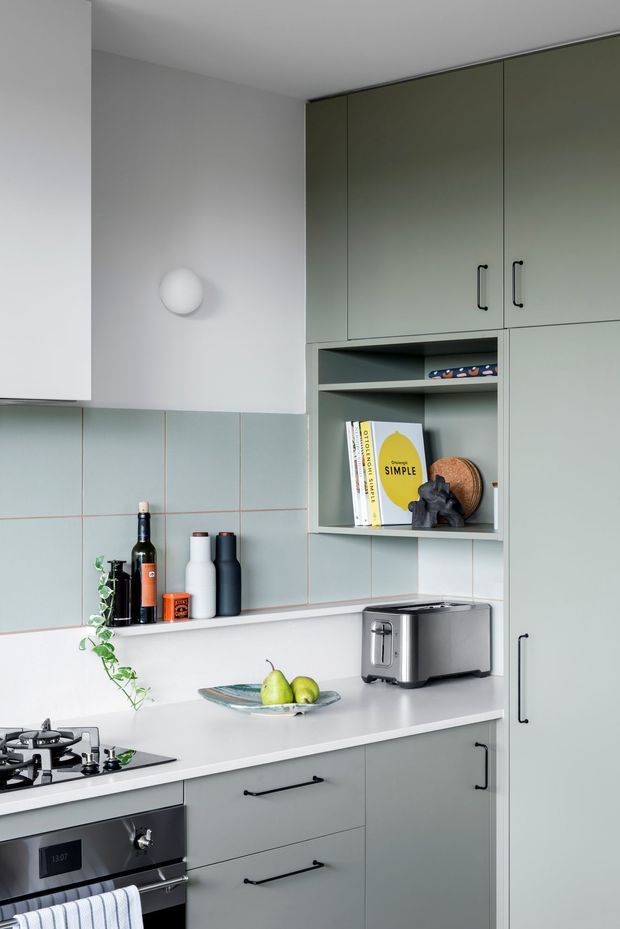 Compact contemporary kitchen with muted sage-green flat-panel cabinetry, pale blue-green tiled splashback, white benchtops, integrated open shelving, and black hardware. A gas cooktop and built-in oven sit beside a toaster and fruit bowl, while simple decor and a trailing plant add a restrained lived-in feel.