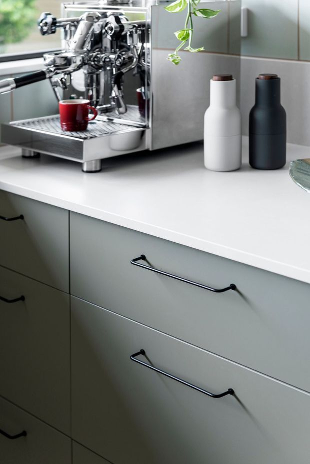 Close-up of a contemporary kitchen coffee station, featuring a polished stainless-steel espresso machine on a white countertop, minimalist white and charcoal bottles, and muted grey-green cabinetry with long black bar handles. A small hanging plant and a red coffee cup add restrained colour accents.