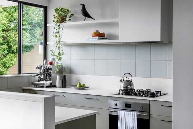 Bright contemporary kitchen with pale grey flat-panel cabinetry, white benchtops, open shelving, and a pale blue-grey tiled splashback. The compact space includes a peninsula, gas cooktop and built-in oven, with black-framed glazing and greenery visible beside the kitchen.