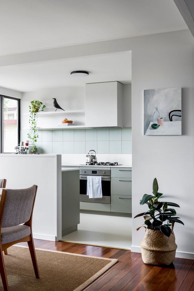 A bright, compact contemporary kitchen viewed from an adjoining dining or living area. The kitchen features pale sage-green lower cabinetry, white upper cabinetry and open shelving, a light blue-green tiled splashback, stainless-steel oven and gas cooktop, and a peninsula dividing it from the dining space. Dark timber flooring continues through the foreground, where a woven rug, upholstered wooden dining chair, potted plant, and minimalist artwork create a warm, refined interior.
