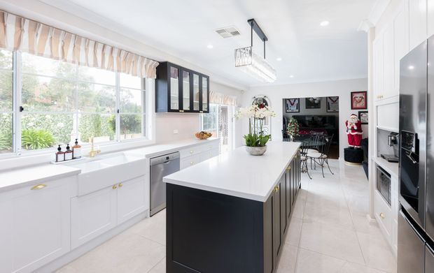 Bright, spacious white kitchen with a large central island featuring a dark charcoal base and white benchtop. The room includes extensive white cabinetry, integrated black stainless appliances, a broad glazed window with striped Roman blind, pendant lighting, and a dining area visible beyond the kitchen.