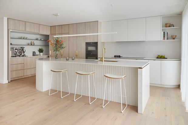 Bright contemporary kitchen with a large fluted island, integrated white cabinetry, pale timber joinery, stone benchtops, open shelving, built-in appliances, and four slim bar stools. The space has a clean, airy appearance with light timber flooring and minimal pendant lighting.