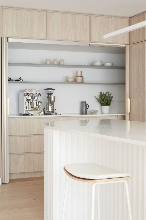 Bright, refined contemporary kitchen with pale timber cabinetry, integrated open shelving, white benchtops and a sculptural fluted island with curved edges. A coffee station with espresso machines occupies the rear bench, while a minimalist white-and-timber stool sits at the island. The palette is soft and neutral with abundant built-in storage and understated decorative objects.