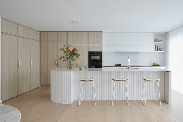 Bright contemporary kitchen with a large white fluted island, three light timber-and-white bar stools, pale timber flooring, and integrated white cabinetry. Full-height light timber cabinetry lines the left wall, while a white minimalist kitchen run with integrated oven, cooktop, sink, and brass-toned tap occupies the rear. Soft daylight enters through sheer curtains on the right.