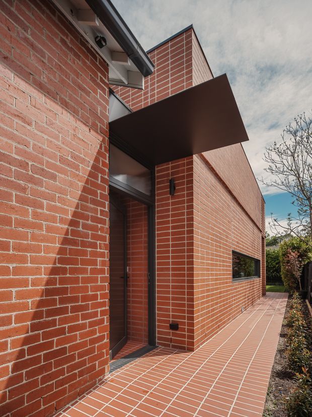 Exterior view of a contemporary brick building focused on the recessed entry and side elevation. Red brick walls and matching brick paving define the facade and narrow walkway, with dark metal-framed glazing, a projecting dark entry canopy, and a simple gabled roof form. Low landscaping borders the path.