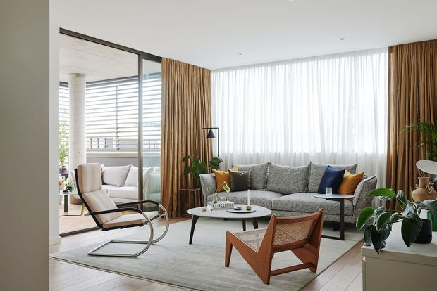 Bright contemporary living room with a large grey sectional sofa, sculptural cantilever lounge chair, wooden and woven leather armchair, round coffee table, indoor plants, and full-height sheer curtains with warm timber drapery. Floor-to-ceiling glazing opens toward a balcony or exterior screened area.