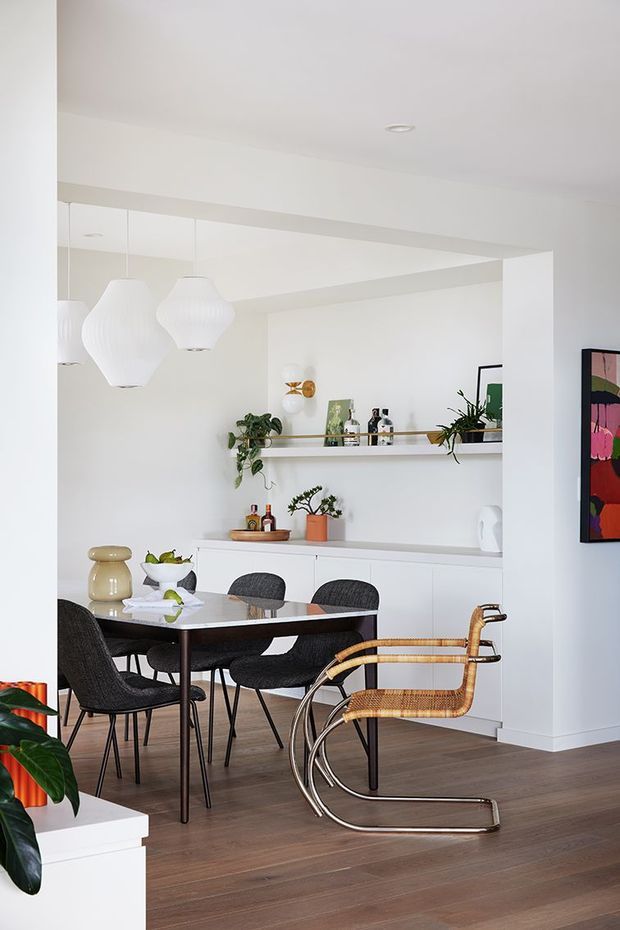 Bright contemporary dining area with a white open-plan interior, dark timber flooring, a rectangular dining table surrounded by woven dark chairs, and a distinctive cantilevered cane-and-metal lounge chair in the foreground. White built-in cabinetry and open shelving display plants, bottles, and decor, while sculptural white pendant lights hang above the table. A colourful framed artwork adds a small accent on the right wall.