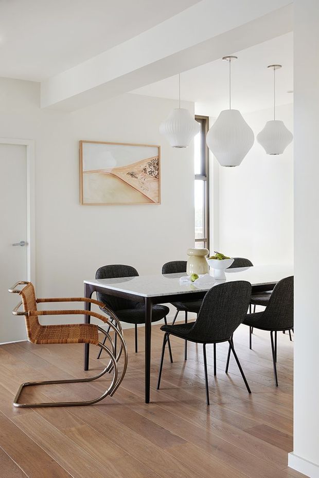 Bright contemporary dining area with a rectangular dark-framed dining table, black upholstered dining chairs, and a distinctive woven rattan-and-chrome lounge chair. Three sculptural white pendant lights hang above the table. The room has white walls and ceiling beams, light timber flooring, minimalist artwork, and a tall window providing natural light.