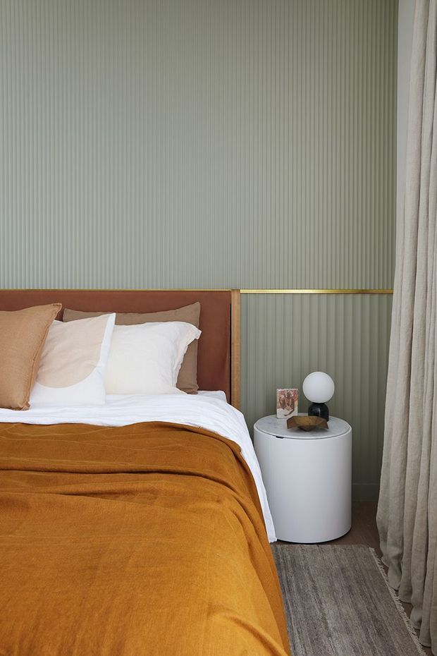 A warmly styled bedroom featuring a bed with a rust-orange velvet coverlet, cream and tan pillows, and a simple upholstered brown headboard. The walls have vertically fluted pale sage-green panelling with a brass-toned horizontal trim. A white cylindrical bedside table holds a spherical lamp and small decor, while full-length beige curtains frame the right side. A grey textured rug adds contrast beside the bed.