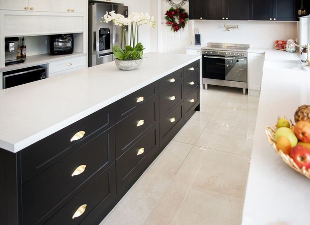Bright, spacious contemporary kitchen featuring a large central island with dark raised-panel cabinetry and brass cup pulls, white stone countertops, extensive white cabinetry, integrated appliances, a freestanding stainless-steel range, and pale large-format tiled flooring. Decorative orchids, flowers, and a fruit bowl add colour, while the overall scheme is crisp and high contrast.