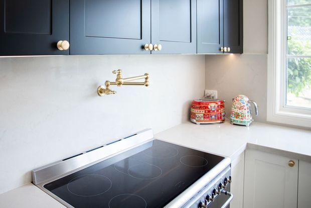 A compact, refined kitchen cooking area with a black electric cooktop and stainless-steel range, pale stone-look benchtops and splashback, deep navy-blue shaker cabinetry, brass knobs and pot-filler tap, and a bright window above light cabinetry.