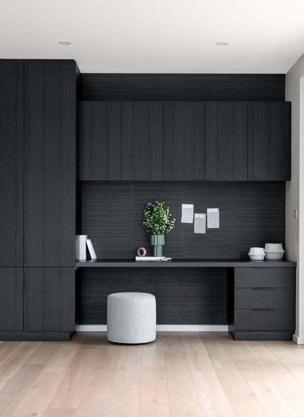 A compact, highly tailored home office with floor-to-ceiling dark timber-look cabinetry, overhead cupboards, a built-in desk, and drawer storage. The dark joinery contrasts with pale timber flooring and white walls, while a light upholstered stool and minimal accessories soften the monochromatic scheme.