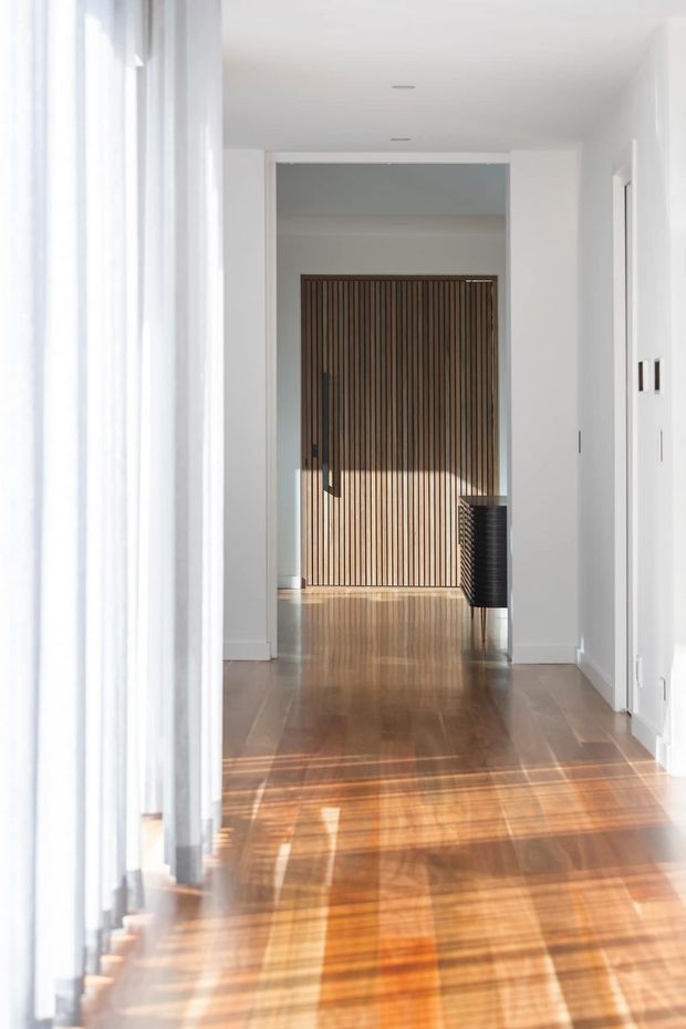 Bright minimalist interior hallway with white walls and ceiling, warm polished timber flooring, sheer full-height curtains along the left side, and a feature wall of vertical dark timber battens at the far end. Strong sunlight creates striped shadows across the floor, with a dark ribbed console or cabinet partially visible near the opening.