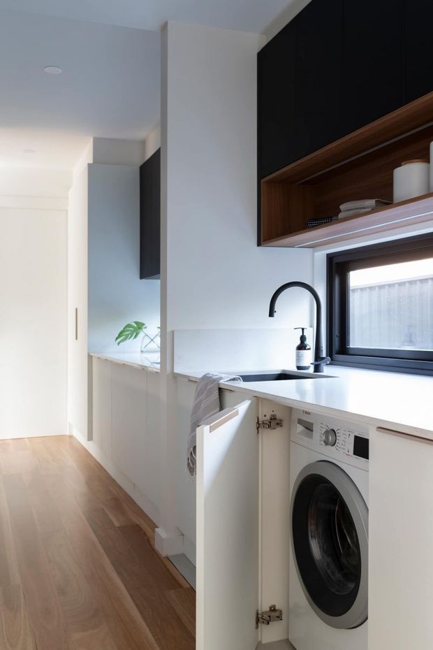 Bright, compact contemporary laundry with a white benchtop, integrated front-loading washing machine, undermount sink and matte black gooseneck tap. White cabinetry and walls are paired with dark cabinetry, warm timber open shelving, a black-framed window and light timber flooring. A hallway and additional cabinetry extend beyond the laundry.