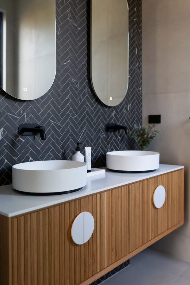 A contemporary bathroom vanity with two round white vessel basins, matte black wall-mounted tapware, and elongated rounded mirrors. The vanity features vertical timber slat cabinetry with large white oval handles and a pale benchtop. A dark grey herringbone tile backsplash creates a strong geometric feature, complemented by neutral plaster walls and grey floor tiles.