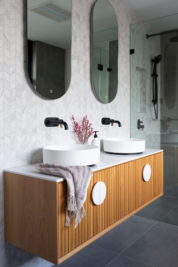 Contemporary bathroom featuring a floating double vanity with ribbed timber cabinetry, white vessel basins, matte black wall-mounted tapware, and two rounded-rectangle mirrors. The walls are finished with white geometric textured tiles, while a glass-enclosed shower with black fittings is visible to the right. Dark grey floor tiles and warm timber joinery create a refined, modern contrast.