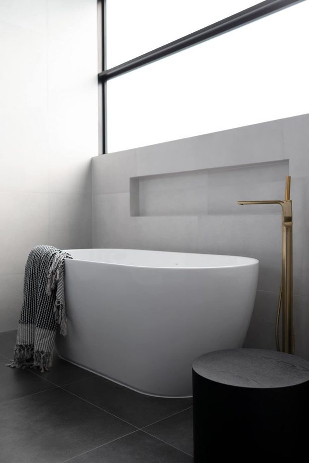 A minimalist contemporary bathroom featuring a freestanding white oval bathtub, dark large-format tiled flooring, pale grey wall tiles with a recessed shelf, and a slim brushed-gold floor-mounted bath tap. A black round stool or side table sits in the foreground, while a textured monochrome throw drapes over the tub. Tall black-framed glazing brings in abundant natural light.