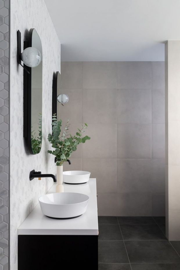 A contemporary bathroom featuring a long white vanity with two round vessel basins, matte black wall-mounted taps, tall rounded mirrors, and spherical wall lights. The walls combine a white textured tile feature wall with large light-grey tiles, while the floor is finished in dark grey tiles. A simple eucalyptus arrangement adds an organic accent.