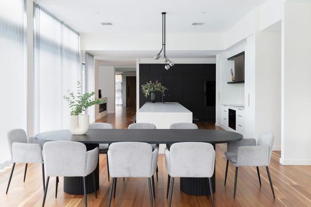 Bright, open-plan contemporary dining and kitchen interior with a long dark timber dining table, upholstered dining chairs, timber flooring, a white island bench, dark kitchen cabinetry, extensive sheer-glazed windows, and a modern pendant light.