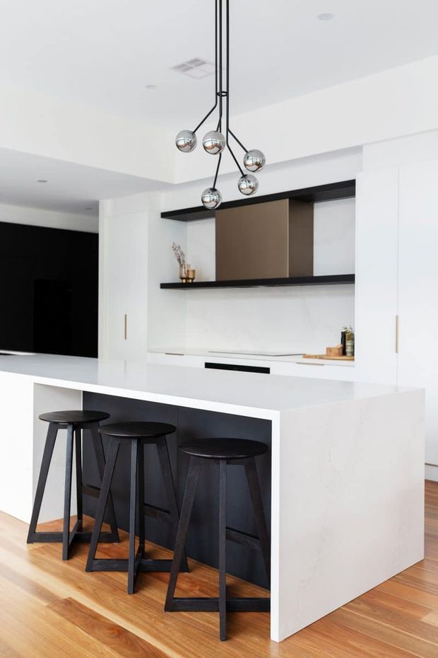 Bright contemporary kitchen featuring a large white island with integrated seating, three black bar stools, white cabinetry, black open shelving, a dark feature panel, warm timber flooring, and a multi-globe pendant light.