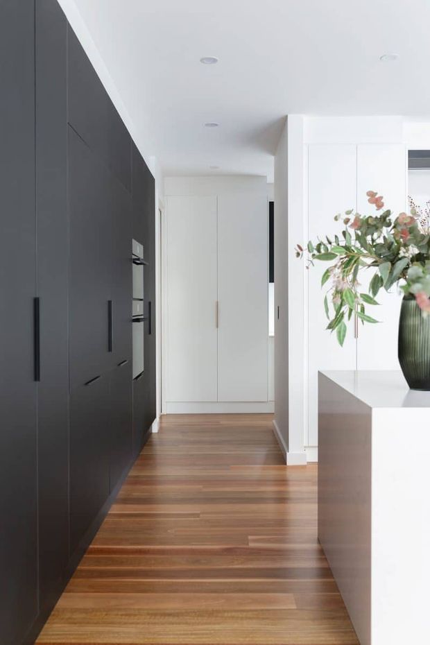A bright, minimalist residential hallway adjoining a kitchen or built-in cabinetry zone. The corridor features full-height matte dark cabinetry with long black handles on the left, white built-in doors and walls at the end, warm timber flooring, recessed ceiling lights, and a white island or console with a dark vase and greenery on the right.