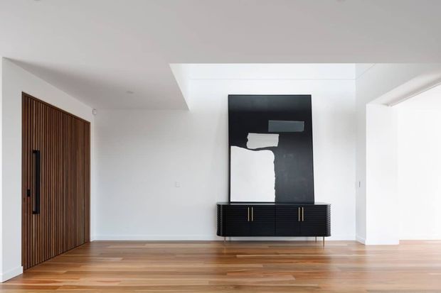 A bright minimalist entry hall with white walls and ceiling, warm timber flooring, a tall vertical timber slat door with a black pull handle, and a low dark built-in console beneath a large abstract black-and-white artwork. A recessed ceiling opening or skylight adds natural light, while a wide opening leads toward another area on the right.