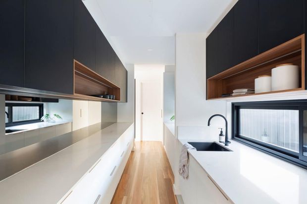 A narrow, highly streamlined galley kitchen with white benchtops and lower cabinetry, dark charcoal overhead cabinetry, and warm timber open shelving. Long horizontal windows bring in daylight, while the timber floor and concealed storage create a clean, contemporary built-in interior.