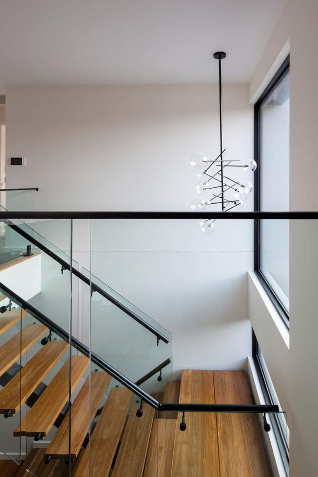 Bright contemporary stairway with warm timber treads and flooring, clear glass balustrades framed in black metal, and a sculptural multi-bulb pendant suspended in a tall stair void. Large vertical glazing brings in abundant natural light, while the white walls and clean detailing create an airy, refined interior.