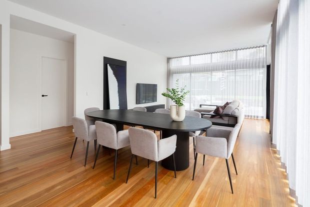 Bright, open-plan dining and living area with a large oval black dining table surrounded by upholstered chairs, warm timber flooring, white walls, floor-to-ceiling sheer curtains, and a compact lounge area beside expansive glazing. A sculptural black pendant is not visible; the space is defined by clean contemporary furnishings and abundant natural light.