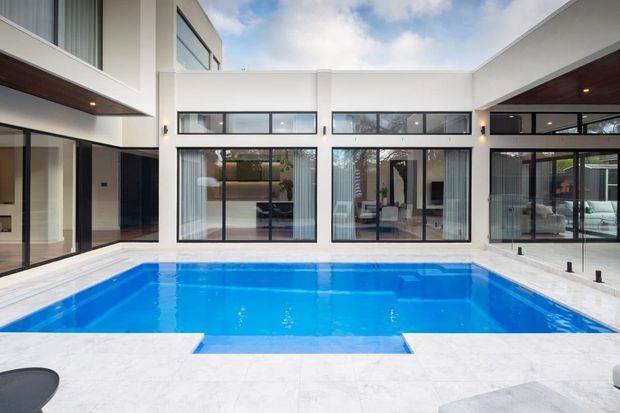 Contemporary two-storey residence arranged around a bright blue swimming pool courtyard. The image shows crisp white rendered walls, extensive black-framed glazing, projecting roof forms, dark timber soffits, and pale stone or concrete pool surrounds. Interior living spaces are visible through the glass, while the pool and courtyard dominate the foreground.
