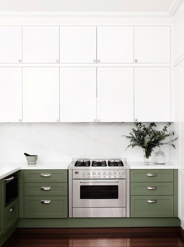 Bright, compact kitchen with deep green lower cabinetry, white upper cabinets, a white veined stone splashback and benchtop, and a stainless-steel freestanding oven and cooktop. Dark timber flooring, polished chrome hardware, and restrained botanical and glass decor create a refined, classic look. The cabinetry extends around a corner, with a tall integrated appliance unit visible at left.